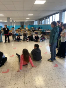 Un groupe d'élèves et un professeur sont rassemblés en cercle, certains assis par terre et d'autres debout, au milieu d'une grande salle de classe ou d'un réfectoire avec des tables et chaises rangées en arrière-plan.